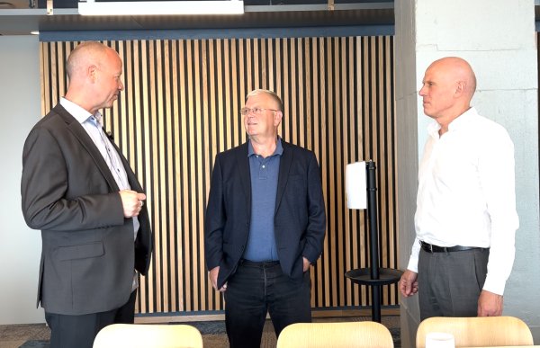 Mann i mørk grå dressjakke og blå skjorte står i åpent kontorlandskap og samtaler med to kollegaer – den ene i hvit skjorte, den andre i mørk dressjakke og mørkeblå skjorte.