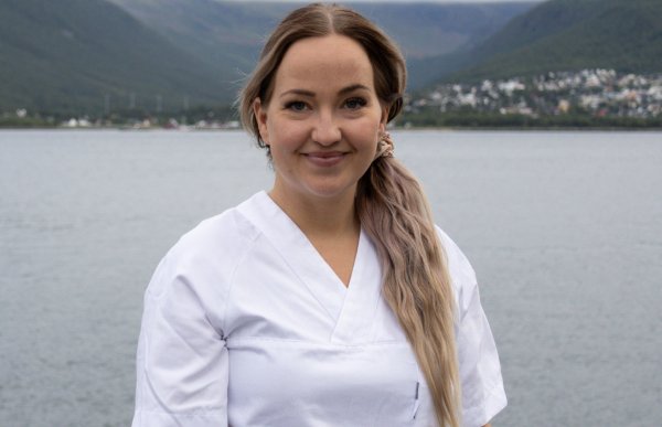 En kvinne med langt og lyst hår. Hun er ikledd hvit helseuniform. Bildet er tatt utendørs, og bak henne er fjell.