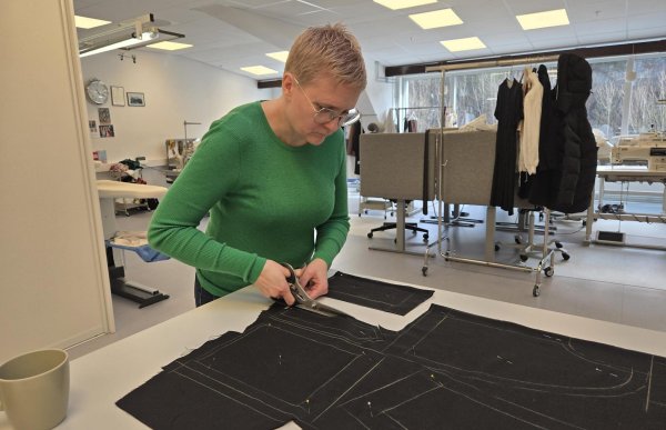 Dame med grønn genser og lyst, kort hår og briller står over et bord og klipper med en saks i et svart stoff.