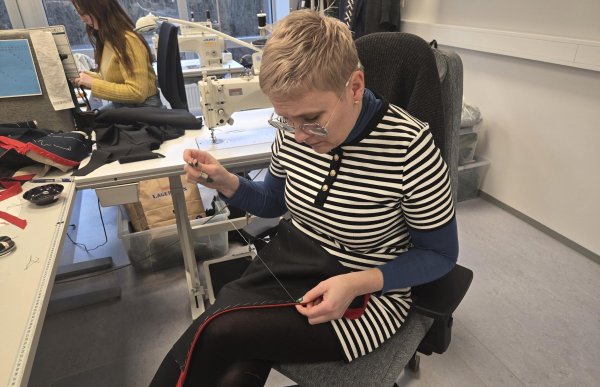 Dame med lyst hår, briller og stripete genser sitter på en kontorstol med nål og tråd og syr i et svart stoffstykke