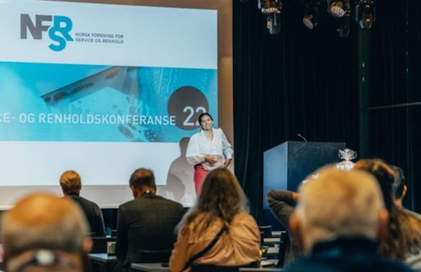 Dame med hvit t-skjorte og brunt hår står på en scene med en presentasjon bak seg. I publikum sitter det flere voksne og hører på.