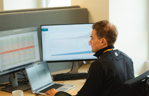 Ung mann i sort genser sitter foran en laptop og to pc-skjermer med ryggen til