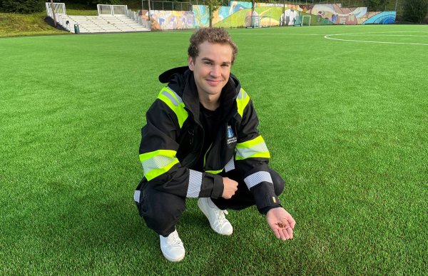 Ung mann i svart og signalgul jakke sitter på huk på en kunstgressbane og viser frem håndflaten med kork. 