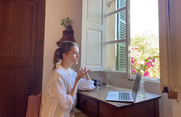 En dame med brunt hår og hvit skjorte sitter ved en pult i et videomøte, pcen står på pulten. Pulten er plassert rett ved et åpent vindu og utenfor er det lyst, med grønne trær og rosa blomster. Bak hun er det en innedør i mørk brun farge, samme farge som på pulten. Det henger også en brun potte på veggen bak henne med en grønn blomst i. 