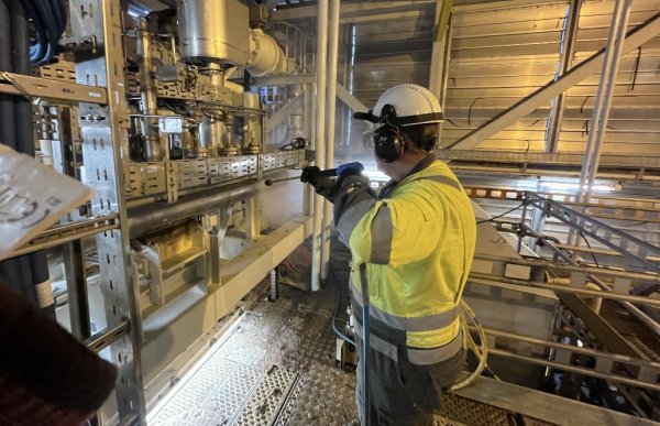 En mann i arbeidsklær med refleks, hjelm og verneutstyr står på en metallplattform og bruker høytrykksspyler mot et metallrør. Området rundt består av rør, metallgitter og strukturer i stål. Det er god belysning, og damp og vannsprut er synlige i luften mens arbeidet pågår.