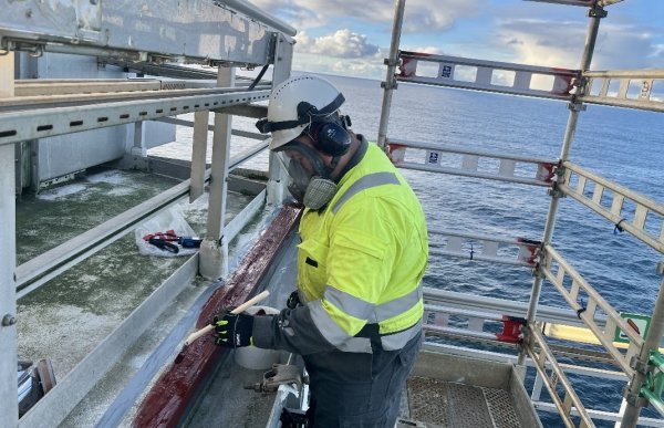 En mann står utendørs på et stillas over havet. Han har på seg en gul vernejakke med refleksstriper, mørke arbeidsbukser, hansker, hjelm og vernemaske. Personen holder en pensel og maler en metallbjelke rødbrun. Rundt ham er det metallrammer og verneutstyr, og i bakgrunnen skimtes et rolig hav under en delvis skyet himmel.