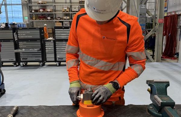 Mann med oransje arbeidsklær, hvit hjelm og hansker står ved et bord og jobber med å sammenstille en kuleventil