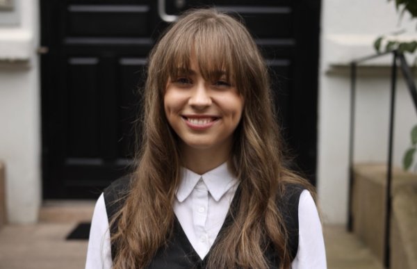 Bildet viser en ung kvinne som står utendørs foran en bygård med mørk inngangsdør. Hun smiler og ser rett mot kameraet. Hun har langt, lysebrunt hår med pannelugg, og hun er kledd i en hvit skjorte med en sort vest utenpå. Bak henne ser man steintrapper og litt grønt løv fra en plante ved siden av trappen.