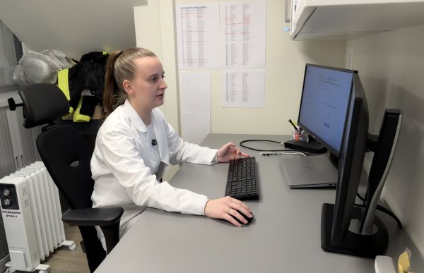 En kvinne sitter ved en kontorpult og jobber på en datamaskin. Hun har håret i hestehale og er kledd i en hvit arbeidsfrakk. På pulten står en skjerm, tastatur og mus, og bak henne henger noen ark på veggen.