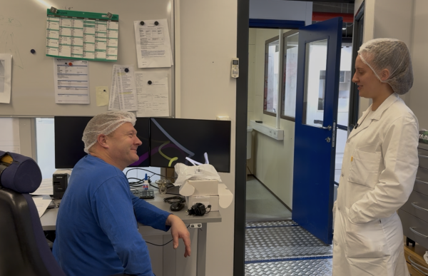En mann og en kvinne er i et arbeidsmiljø som ser ut som et produksjons- eller laboratorieområde. Begge har på seg hårnett, og kvinnen har i tillegg hvit arbeidsfrakk. Mannen sitter ved en pult med skjermer og utstyr, mens kvinnen står og snakker med ham. I bakgrunnen er det oppslagstavle med papirer og en åpen dør inn til et annet rom.