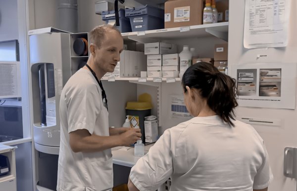 En mannlig og en kvinnelig helsefagarbeider i hvite sykepleieruniformer står foran en hylle med medisiner