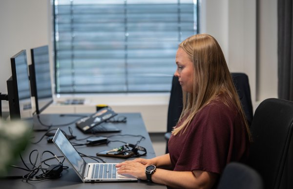 Kvinne med langt blondt hår sitter ved et skrivebord og skriver på en bærbar PC. På skrivebordet står flere skjermer og annet datautstyr.