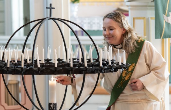 Kvinne med skulderlangt lyst hår i hvit liturgisk drakt og grønn stola tenner et lys på en stor lysglobe i en kirke.