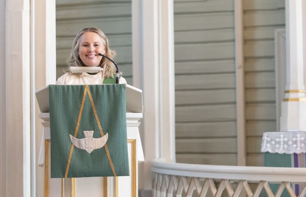 Kvinne med lyst skulderlangt hår står bak en talerstol i en kirke. Hun smiler og har en mikrofon foran ansiktet. Talerstolen er dekorert med et grønt klede med et hvitt symbol av en fugl.