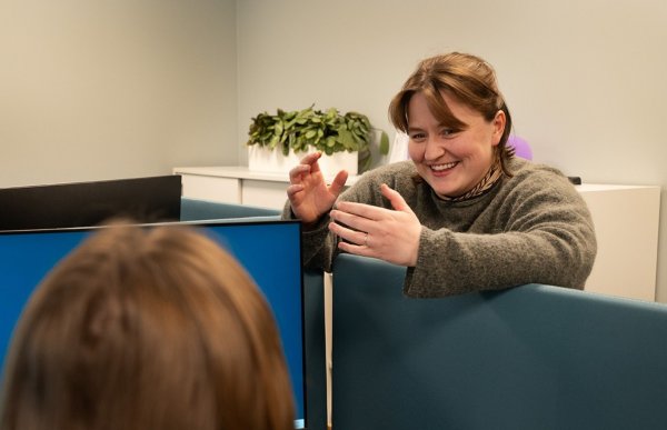 Ung smilende kvinne med brunt halvlangt hår og beige genser, er i samtale med en annen kvinne. Den andre kvinnen sees bakfra og har også brunt hår. De befinner seg i et kontorlandskap. 