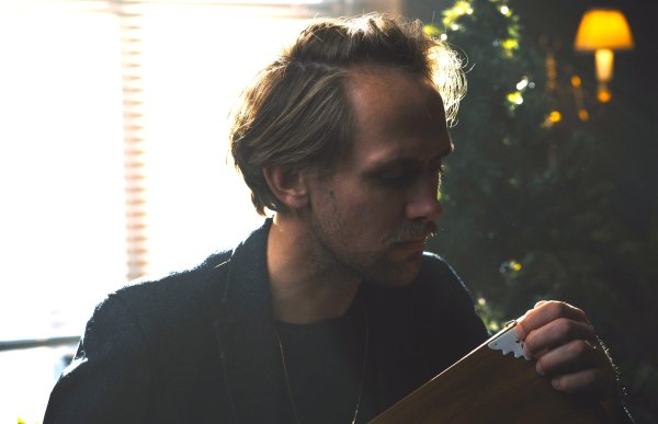 En mann med mørk dressjakke sitter i et rom foran et juletre.