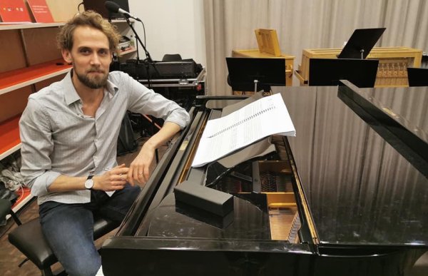 En ung mann i dongeribukse og lys skjorte, sitter foran et piano med noteark.