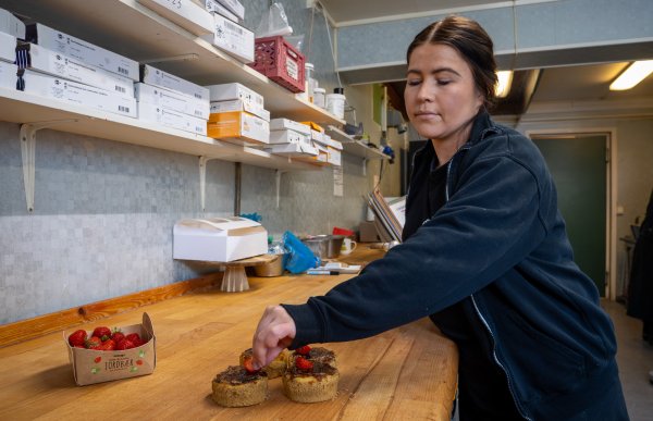 En kvinne i svart jakke står ved en benk og pynter små kaker med bringebær.