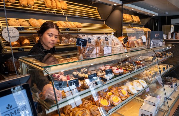 En kvinne står bak en disk fylt med bakevarer og kaker inne på et bakeri. Kvinnen setter prislapper foran varene. Bak henne er det brød i brødhyllene.