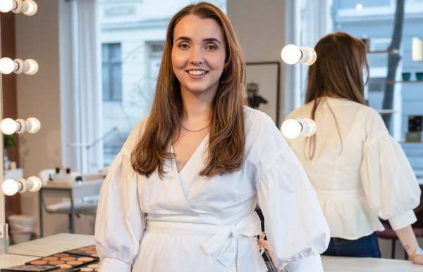 Kvinne i hvit bluse står foran stort sminkebord med speil og smiler til kamera.