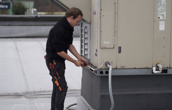 En mann står og skrur på en stor ventilasjonsmaskin på et tak. Han har på seg en sort fleece og en sort arbeidsbukse med verktøy i. 