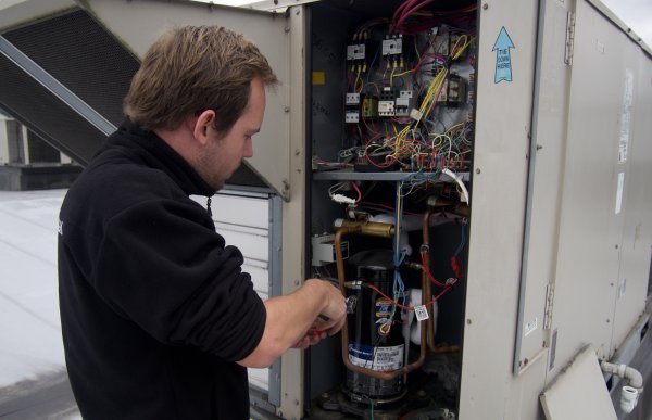 En mann står foran et elektrisk skap med masse koblinger, rør og ledninger. Bildet er tatt bakfra, så man ser inn i hele skapet. Han har en tang i hendene og skrur på et tynt rør inne i skapet. 