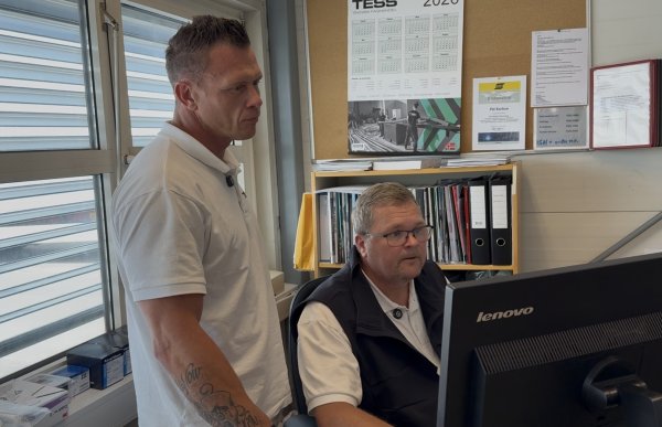 En mann med kort, mørkt hår og tatoverte armer står ved siden av en kollega foran en dataskjerm. Begge har på seg hvite TESS-poloskjorter, mens den sittende kollegaen i tillegg har en mørk vest. De ser begge mot skjermen.