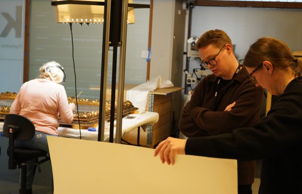 To personer står ved et bord og ser på et stort ark, mens de diskuterer noe sammen. I bakgrunnen sitter en tredje person og arbeider med et kunstobjekt under en lampe i et verkstedmiljø.