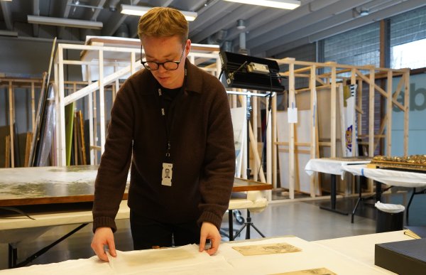 En person står ved et bord og håndterer et ark eller en tynn plate med forsiktighet. På bordet ligger flere papirark med tegninger, og rommet ser ut som et verksted eller arbeidsområde for kunst.
