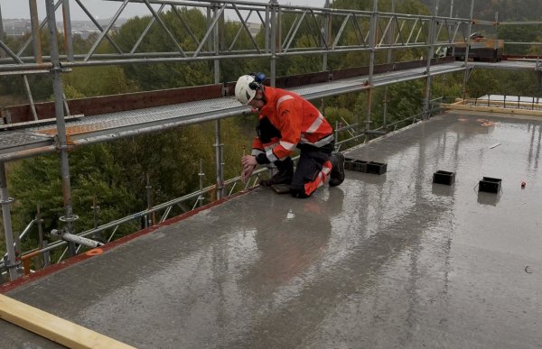 En mann iført en hvit vernehjelm og oransje arbeidsklær med refleks sitter på kne på et vått betongdekke på en byggeplass. Han holder et måleinstrument ved kanten av dekket. Rundt byggeplassen er det stillaser, og nedenfor ses trær og bebyggelse.
