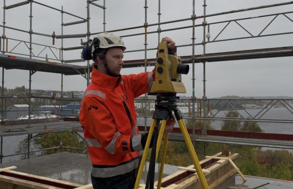 En mann i hvit vernehjelm og oransje arbeidsjakke med refleks står ved et instrument på et gult stativ og justerer det med hendene. Rundt ham er det stillaser, med bebyggelse, trær og vann i bakgrunnen.