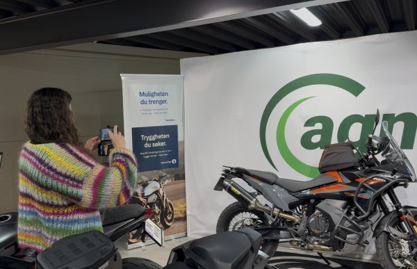 En kvinne med langt, brunt, krøllete hår tar bilde av en motorsykkel med mobilen sin inne i en butikk eller utstillingslokale. Hun har på seg en fargerik, stripete cardigan. Rundt henne står flere motorsykler og reklameskilt.