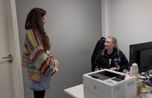 To kvinner snakker sammen på et kontor. Den ene står og har på seg en fargerik, stripete cardigan og et blått olaskjørt. Hun har langt, brunt, krøllete hår. Den andre kvinnen har blondt hår i fletter og svarte klær. Hun sitter ved en skrivepult med dataskjerm og printer. De smiler mot hverandre.