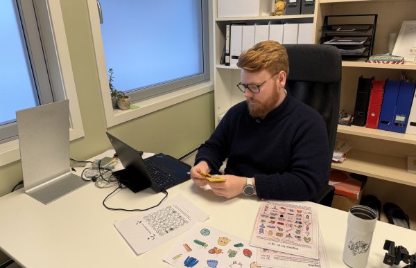 En mann med briller og rødt hår og skjegg sitter ved et skrivebord på et kontor. Han holder gule kort i hendene og ser ned på dem. På bordet ligger en åpen laptop, ark med illustrasjoner, annet treningsmateriell og en kopp. I bakgrunnen er det vindu og hyller med permer og bøker.