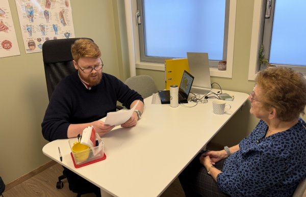 En mann med briller og rødt hår og skjegg sitter ved et bord sammen med en eldre kvinne i et behandlingsrom. Mannen holder et ark og ser ned på det, mens kvinnen sitter på motsatt side og lytter. På bordet ligger ulike hjelpemidler og papirer, og på veggen bak dem henger en plakat med illustrasjoner av hals og strupe.