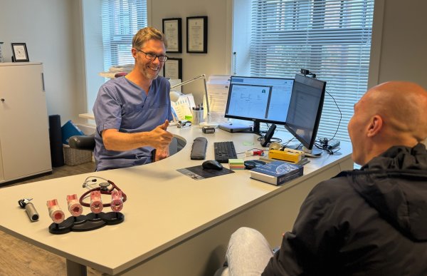Mann i blå skjorte sitter bak et skrivebord med to pc-skjermer. Han har briller og viser en tommel opp til en person som sitter på andre siden av pulten. 