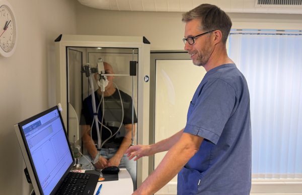 Mann med blå t-skjorte står foran en pc-sjerm. I en glassboks i et hjørne sitter en person med utstyr på munnen for å puste i.