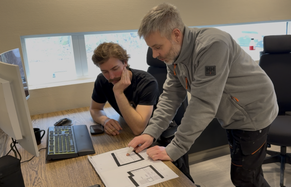 To menn står ved et skrivebord og ser på arbeidstegninger. Den ene peker på tegningen mens de diskuterer løsningen. På bordet ligger tegninger, en datamaskin og en mobil.