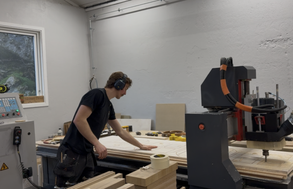 En mann arbeider ved en stor CNC-maskin i et verksted. Han har på hørselsvern og justerer en stor treplate på maskinbordet. Rundt ham ligger treplater og verktøy.