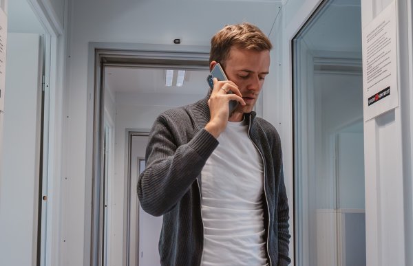 En mann går i gangen på en brakke og snakker i telefonen. Han er kledd i olabukser, hvit t-skjorte og en grå genser.