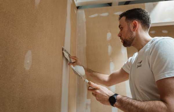 En mann med brunt hår og hvit t-skjorte ser konsentrert på en vegg, mens han sparkler ned langs en skjøt mellom to veggplater.