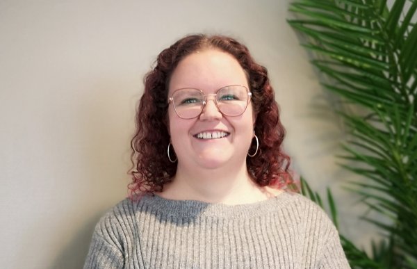 En kvinne med rødlig, krøllete hår og briller smiler mot kameraet. Hun har på seg en grå ullgenser. I bakgrunnen ser vi en plante.