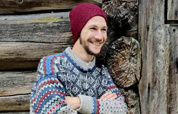 Mann med tradisjonell ullgsenser og rød lue lener seg mot en vegg av tømmer