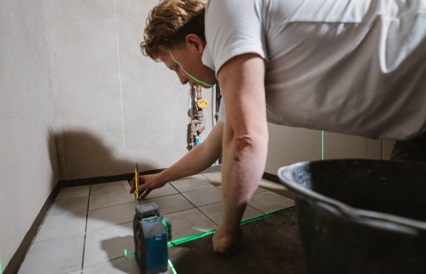 En mann sitter bøyd over nylagte fliser på et gulv. Foran ham står en krysslaser som sender grønne, rette laserlinjer i to retninger. Mannen sjekker avstand mellom flisene med baksiden av en kniv.