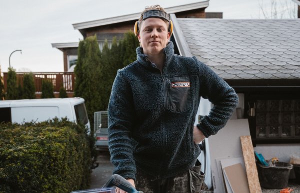 En mann står utenfor et hus med hånda på hofta. Han har på seg en tjukk genser, og hørselvern på hodet. I forgrunnen er et verktøybord med verktøy og materialer.