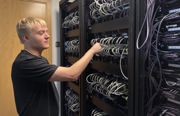 Lyshåret mann i svart t-skjorte står foran et skap med mange hyller hvor det er mange ledninger