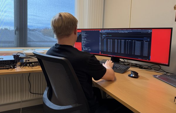 Lyshåret mann med svart t-skjorte sitter foran en bred pc-skjerm som står på en pult. Det er mange tall og mye tekst på prorgrammet på pc-en