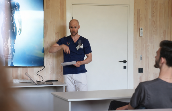 Mann med blå skjorte og hvit bukse står ved en storskjerm og snakker til personer som sitter bak pulter. Han holder en presentasjon i et undervisningsrom. I høyre del av bildet sitter en mann med brunt hår og hører på.