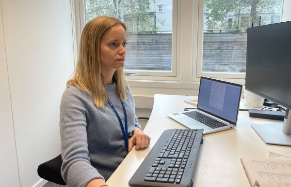 Dame med lyst, halvlangt hår sitter på et kontor. På pulten er det en stor pc-skjerm, tastatur og en liten, bærbar pc. Hun leser noe på skjermen. 