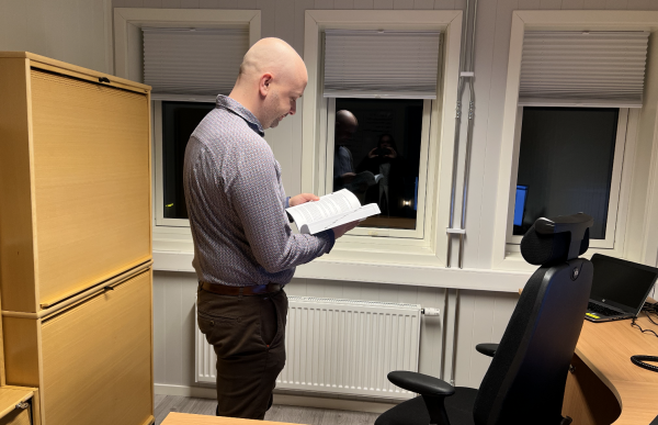 En mann i en lys mønstret skjorte står på et kontor og leser i ei bok. Ved siden av han er en kontorstol og et skrivebord med en bærbar PC på. På den andre siden er et skap.